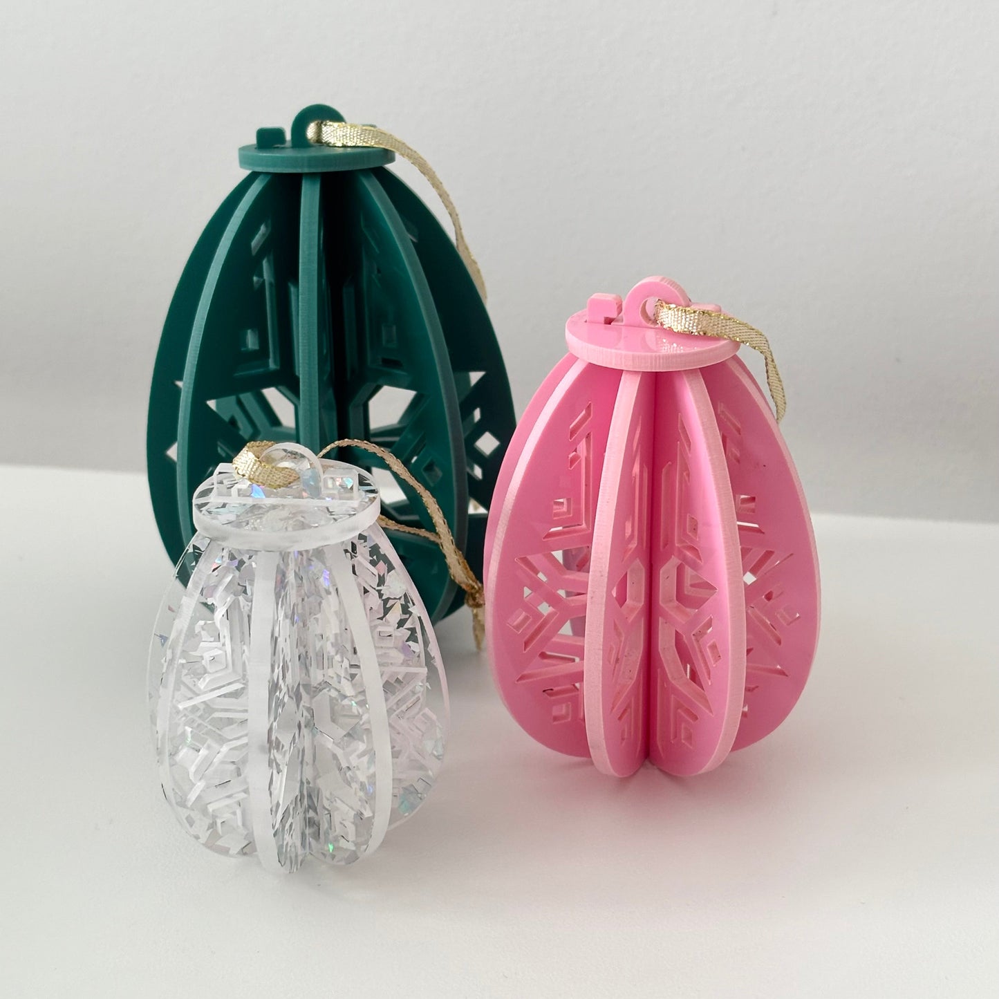 Three decorative easter egg ornaments in green, clear, and pink on a white surface with a light gray background.