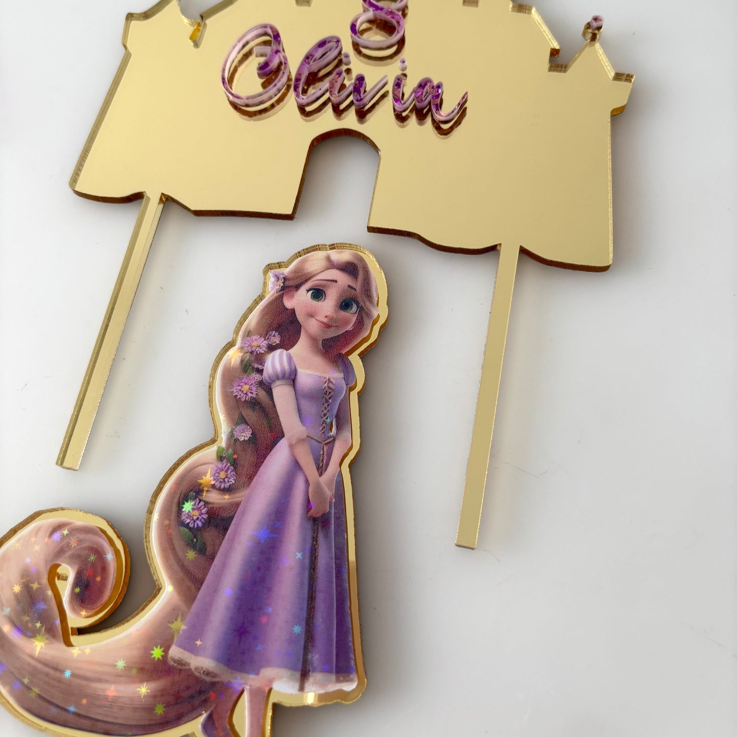 Decorative cake topper with gold castle, Rapunzel figure, and 'Olivia' text on a white background