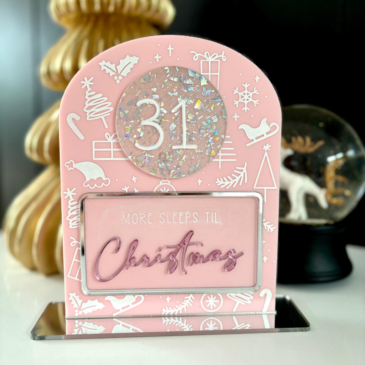Pink Christmas countdown sign with '31 more sleeps til Christmas' text, gold tree in the background.