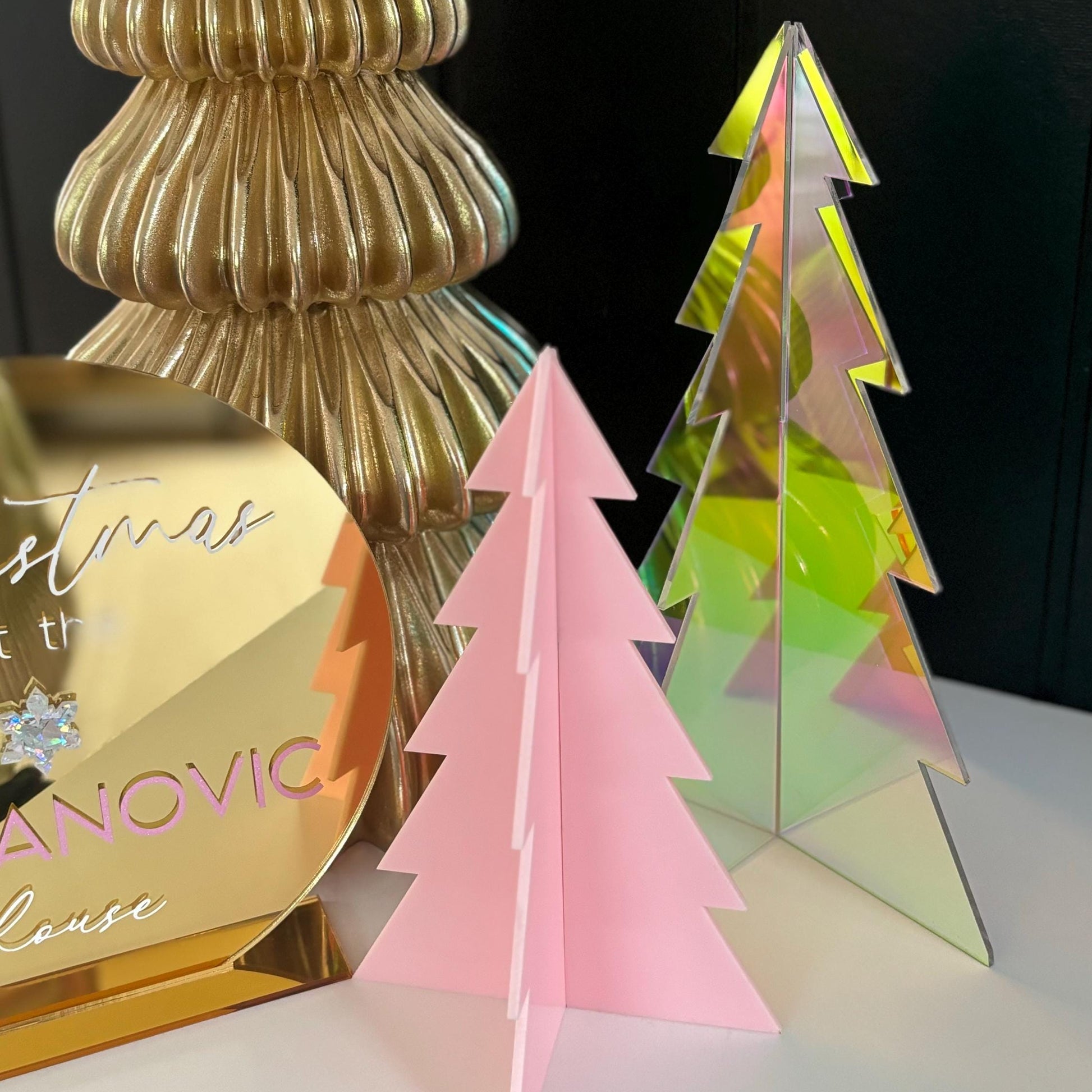 Iridescent and pink Christmas tree-shaped objects on a reflective surface with a gold decorative item in the background.