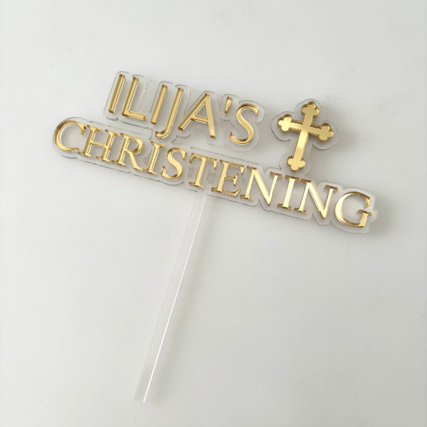 Gold cake topper with 'Ilija's Christening' text and cross on a white background