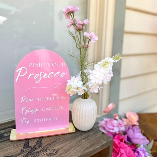 Pink sign with text on a stand next to a vase with flowers on a wooden surface.
