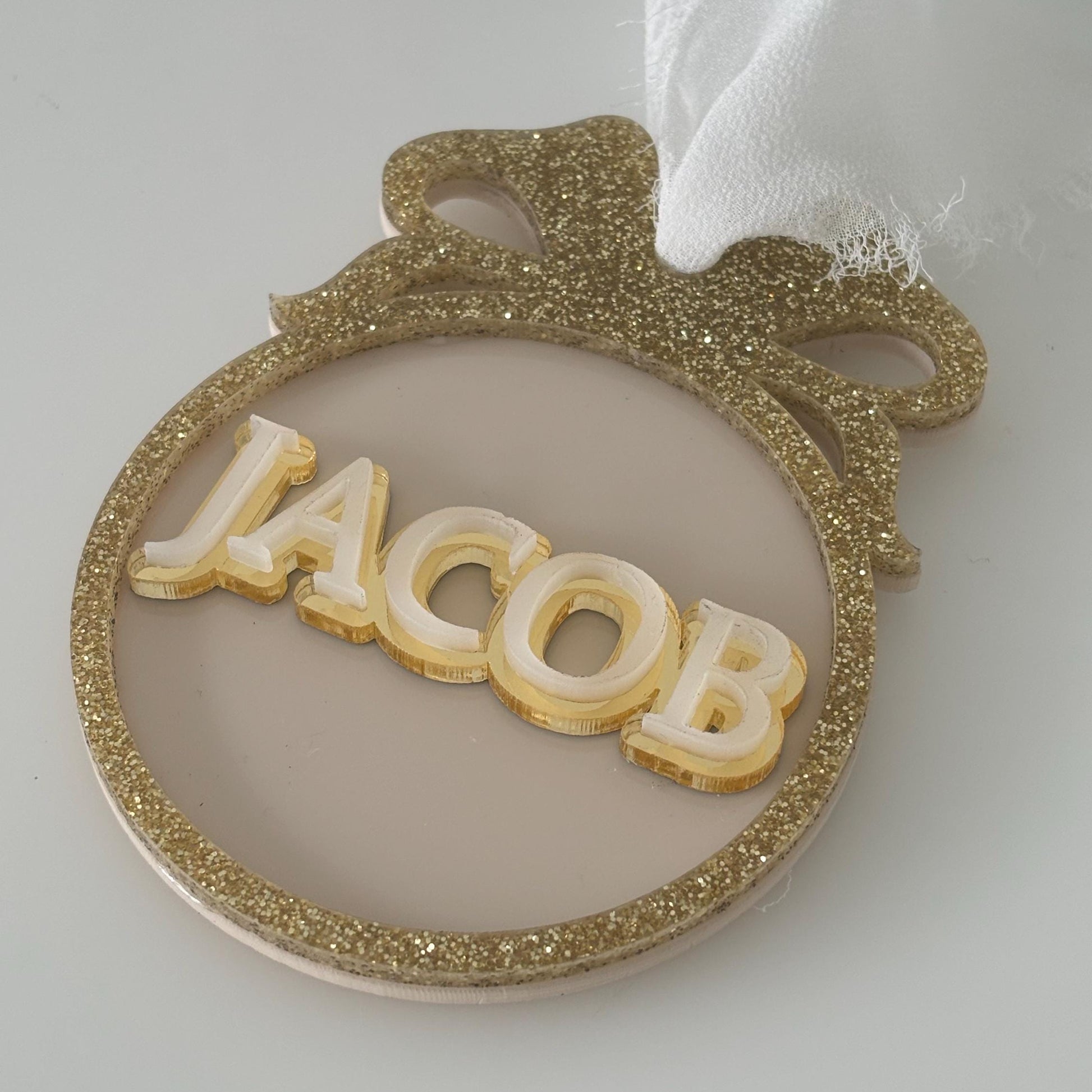 Decorative Christmas bauble with 'JACOB' on a gold glittery frame against a white background