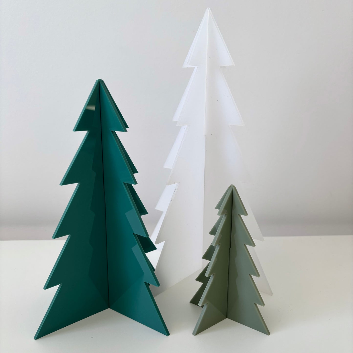 Three acrylic Christmas tree decorations in green, white, and olive green on a white surface with a light grey background.