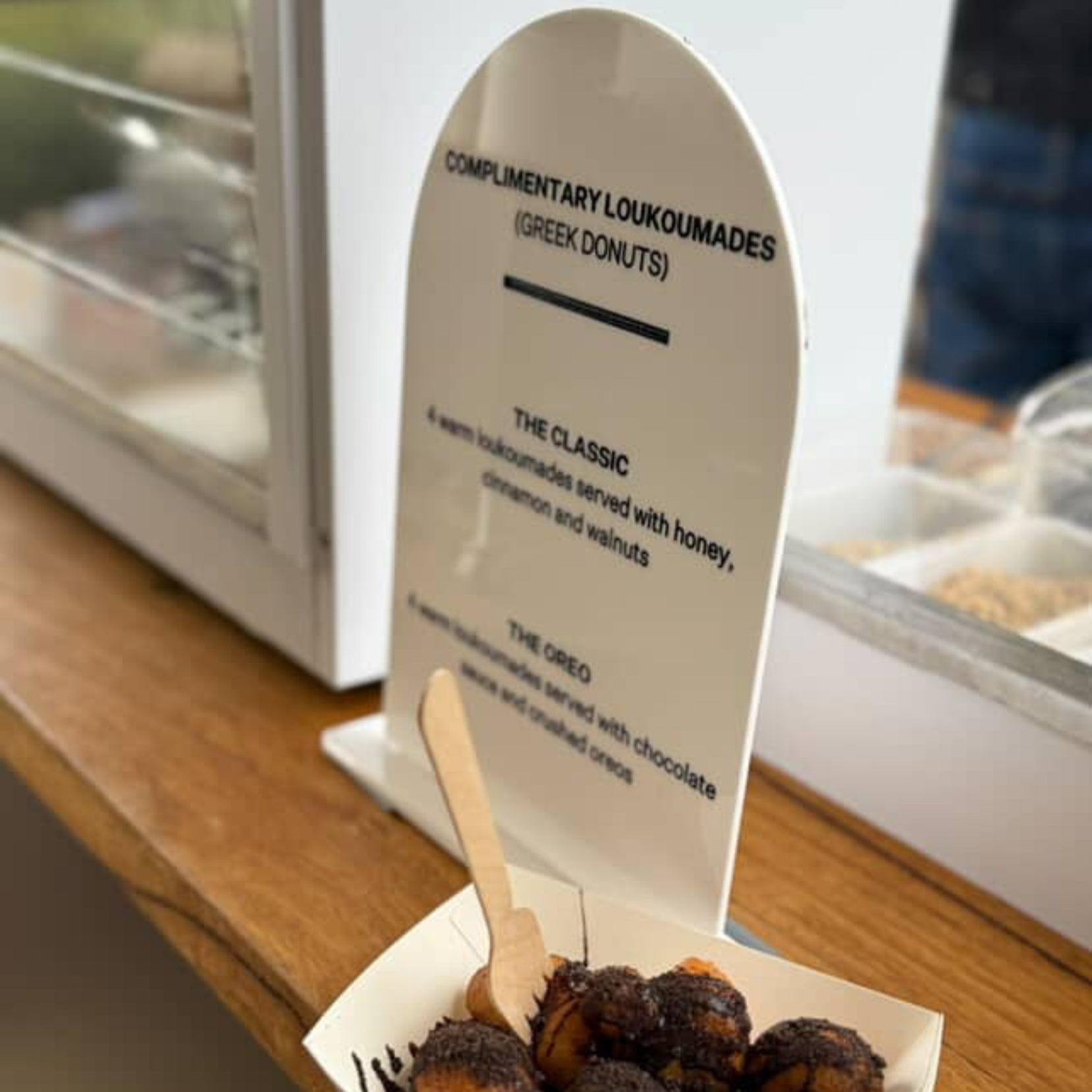 A4 White Sign with Complimentary loukoumades (Greek donuts) on a wooden counter.
