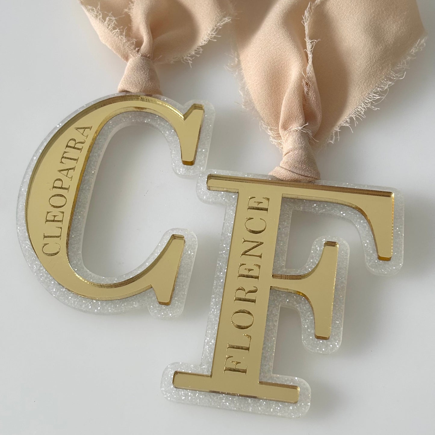 Gold letter 'C' with 'Cleopatra' and gold letter 'F' with 'Florence' on a beige ribbon against a white background.