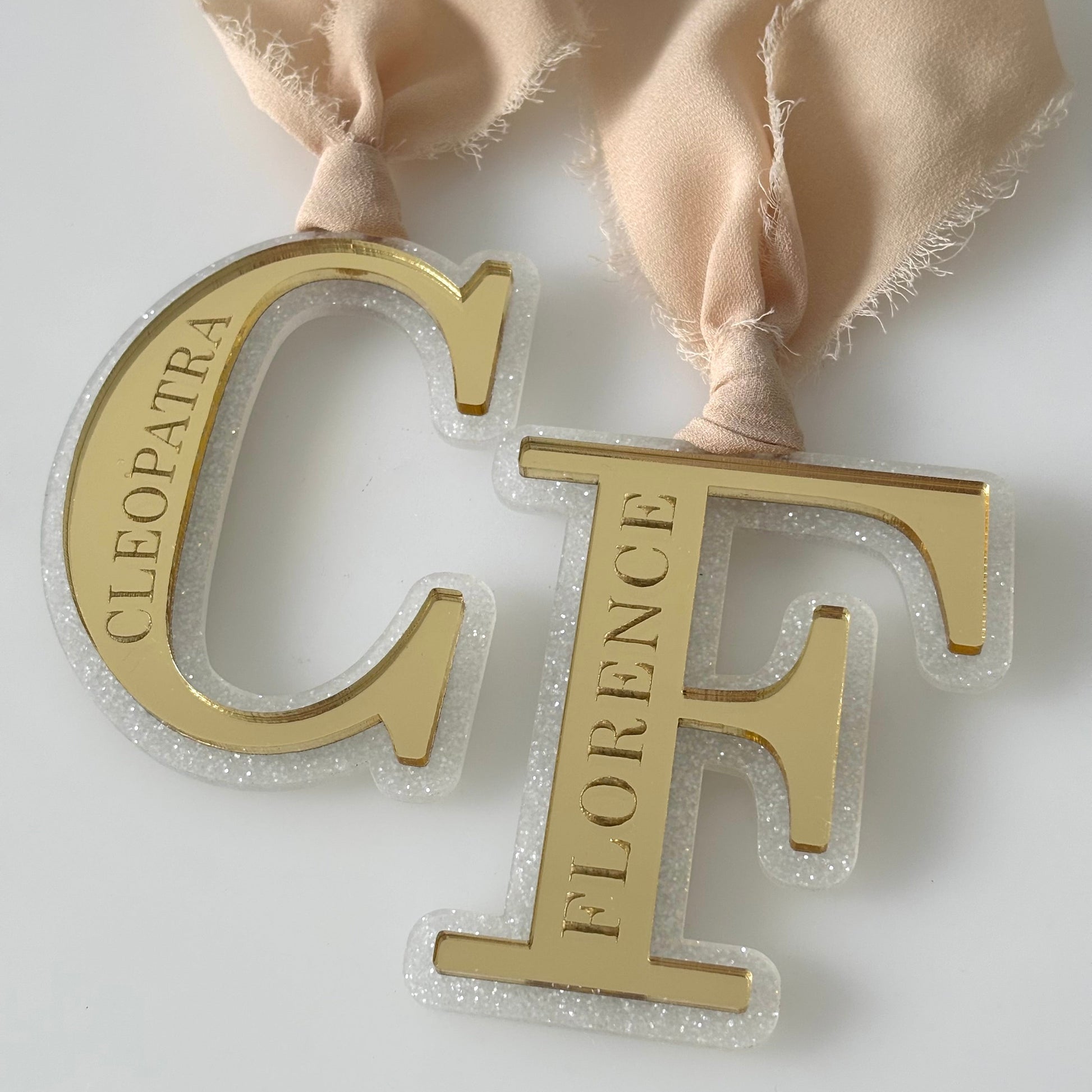 Gold letter 'C' with 'Cleopatra' and gold letter 'F' with 'Florence' on a beige ribbon against a white background.