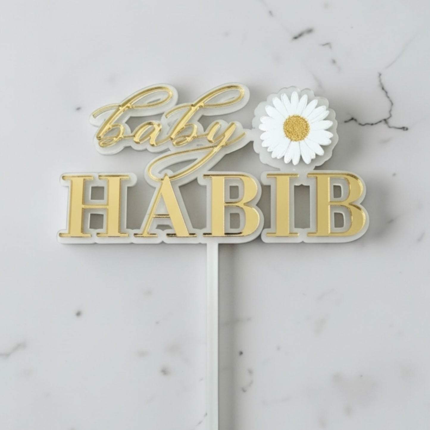 Decorative cake topper with 'Baby Habib' text and a white flower on a marble background