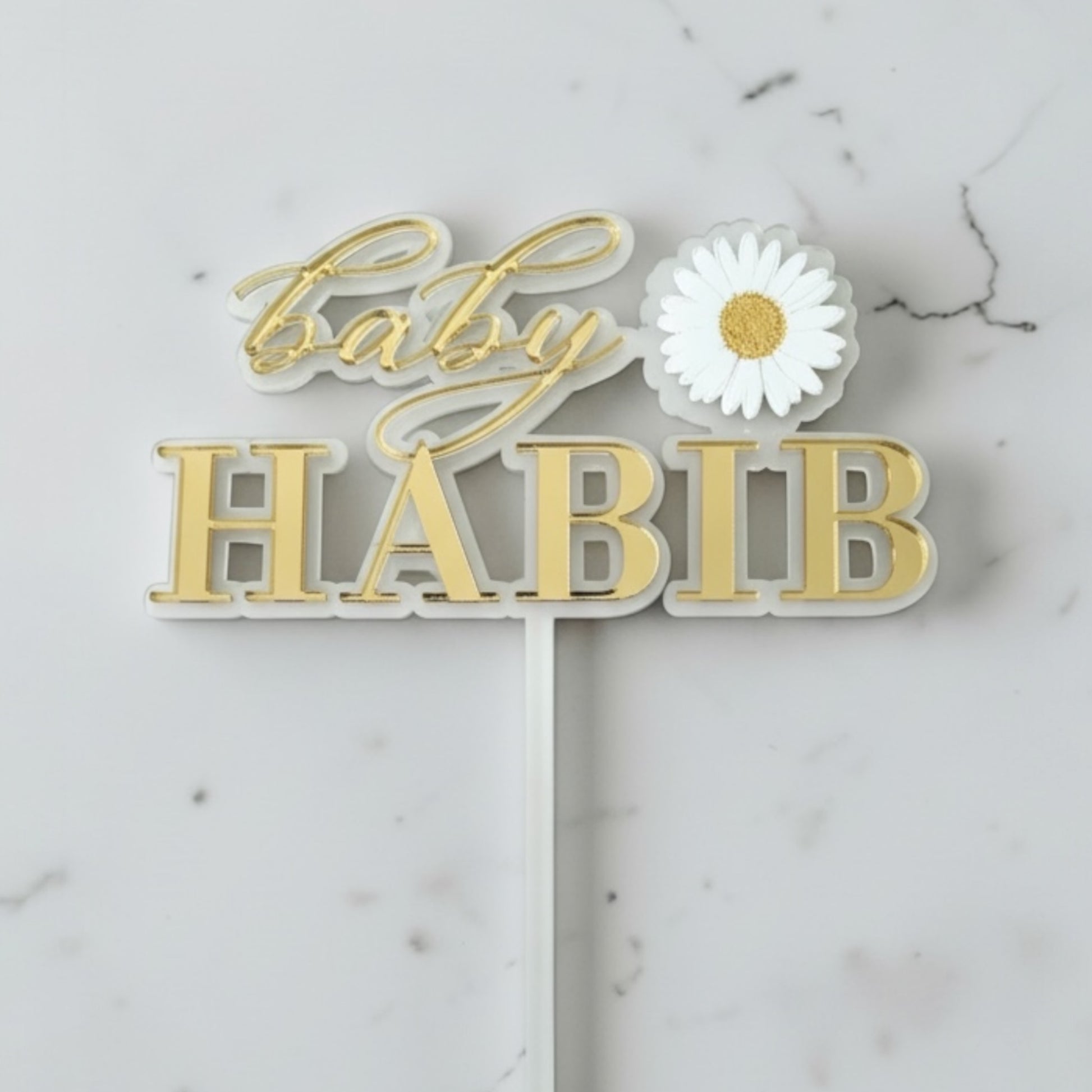 Decorative cake topper with 'Baby Habib' text and a white flower on a marble background