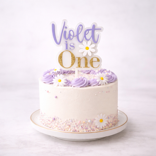White cake with purple frosting and 'Violet is One' topper on a white background