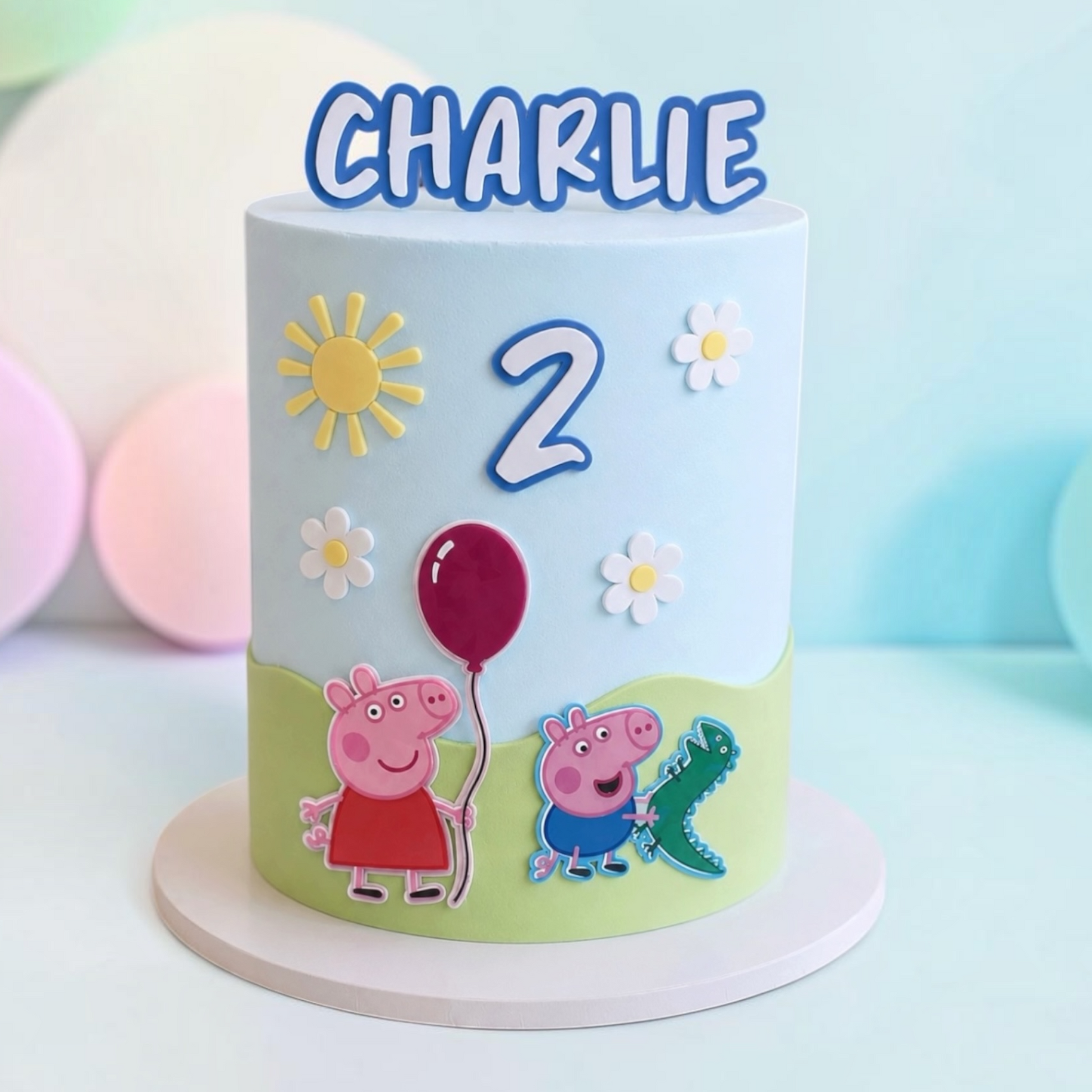 A custom Peppa Pig themed birthday cake decorated with fondant, featuring the name 'CHARLIE' in blue letters on top, a large number '2', Peppa holding a red balloon, and George with a green dinosaur toy. The cake is decorated with a light blue and light green color scheme and is displayed on a white round cake stand.