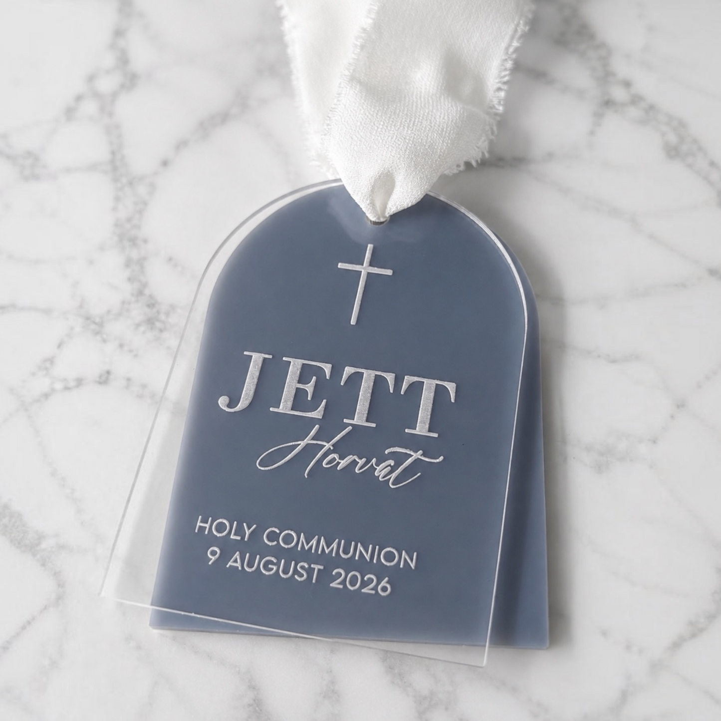 A personalized Holy Communion communion rite tag shaped like a tombstone, printed with a cross and the name 'JETT Horvat' with the date '9 August 2026'. The tag is enclosed in a clear plastic sleeve and attached to a white fabric ribbon, resting on a white and gray marble surface.