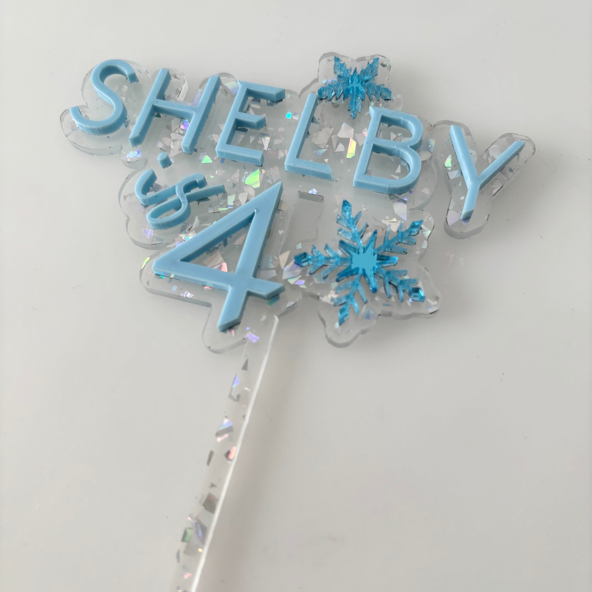 Blue frozen birthday cake topper with 'SHELBY' and number '4' on a white background