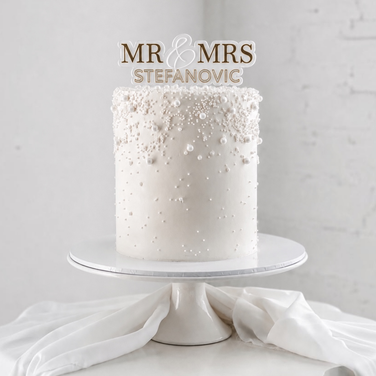 A white frosted cake with decorative sugar pearls sits on a white cake stand against a light gray brick wall. A custom topper reads 'MR & MRS STEFANOVIC' in gold and white lettering.