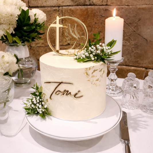 White cake with gold topper and 'Tomi' inscription on a table with flowers and a candle.