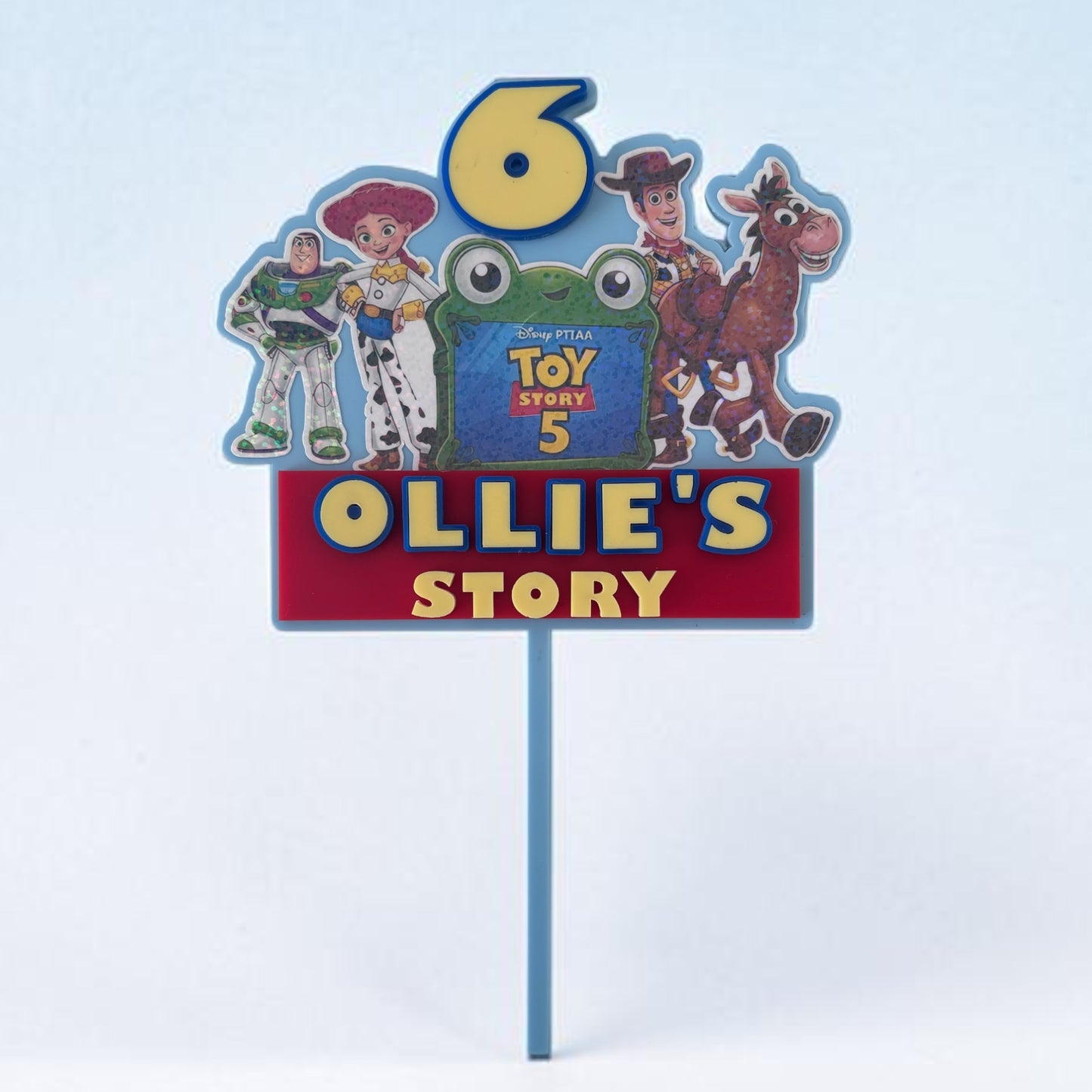 A custom Toy Story-themed birthday cake topper on a blue plastic stake. The sign features a large yellow number 6, characters Woody, Buzz Lightyear, Jessie, and Bullseye, a green frog character, and a red banner reading 'OLLIE'S STORY' in yellow letters.
