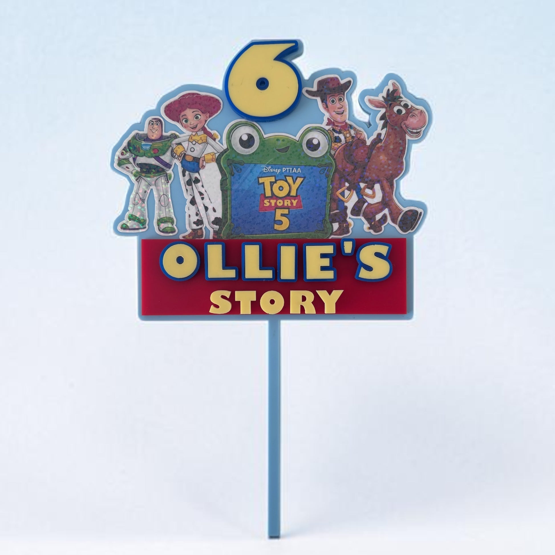 A custom Toy Story-themed birthday cake topper on a blue plastic stake. The sign features a large yellow number 6, characters Woody, Buzz Lightyear, Jessie, and Bullseye, a green frog character, and a red banner reading 'OLLIE'S STORY' in yellow letters.
