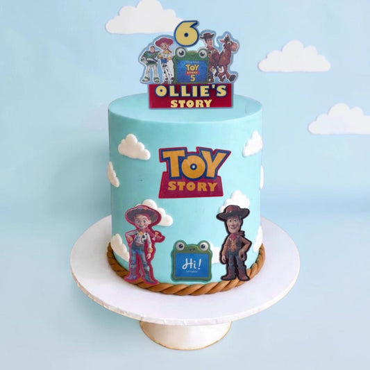 A custom-frosted light blue birthday cake with Toy Story characters and logos, displayed on a white cake stand against a light blue background with white cloud cutouts.